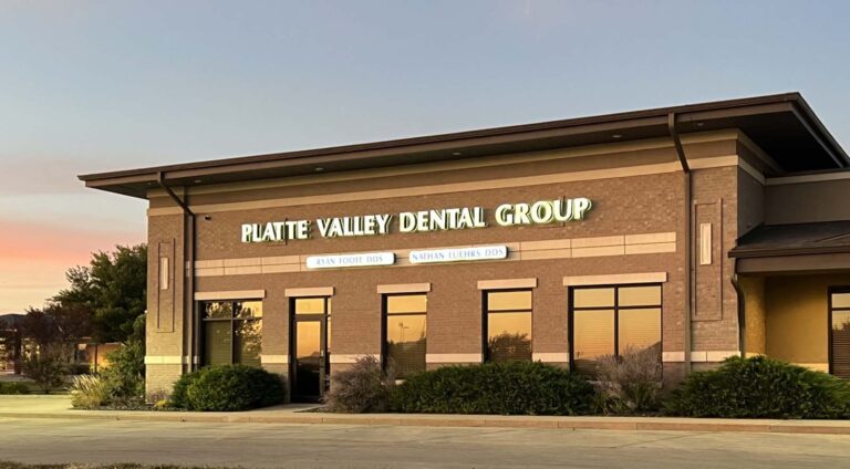 The image depicts the exterior of a building with a sign reading "PLATTE VALLEY DENTAL GROUP." Below the main sign, two smaller signs display the names "RYAN FOOTE DDS" and "NATHAN LUEHRS DDS." The building features a brick facade, large windows, and landscaping with bushes and plants in front.